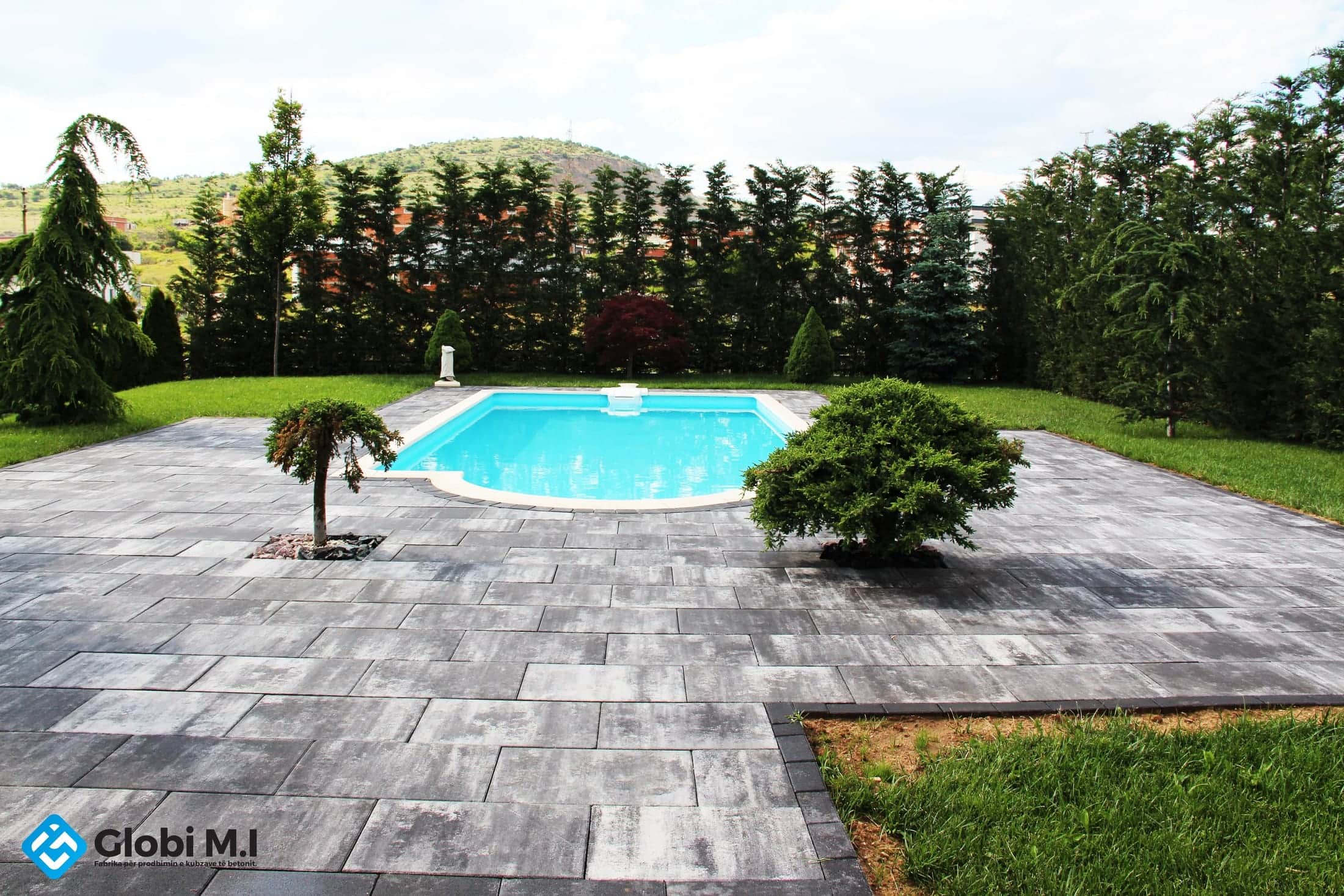 Pool with paving all round, lawn, a conifer hedge and hills behind.