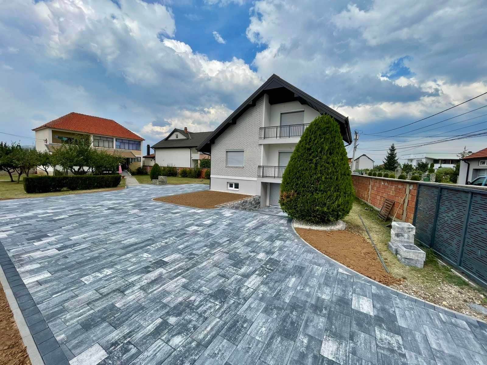 Paved surface running past a house, with lawn and shrubs alongside.