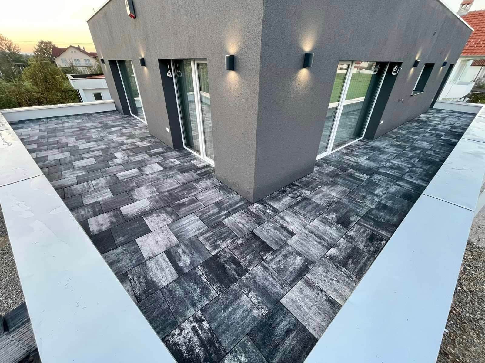 Corner of a grey house at dusk, wall lights on, the paving running round it.
