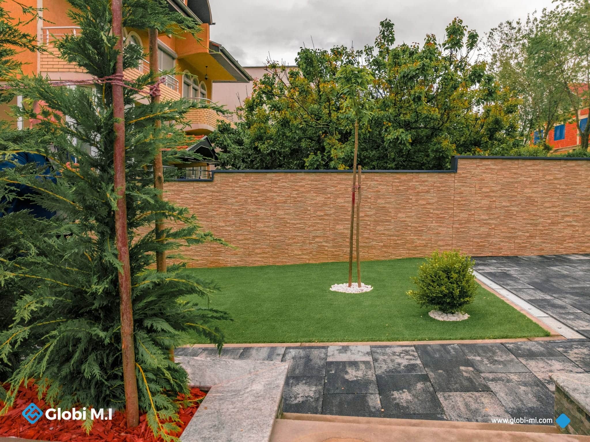 Lawn, trees and a brick wall beside a paved path in a private outdoor space.